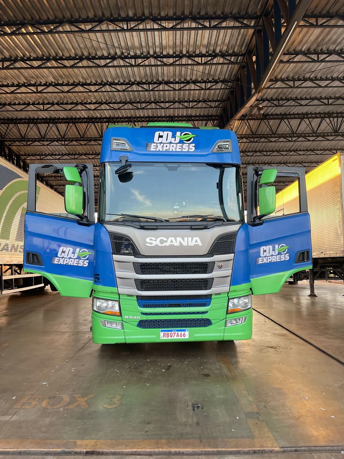 Caminhão azul e verde da CDJ Express visto de frente no galpão